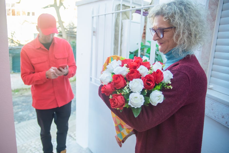 A woman with curly gray hair, wearing glasses and a burgundy sweater, holds a vibrant bouquet of fresh red and white roses wrapped in patterned yellow paper, smiling as she receives the floral arrange