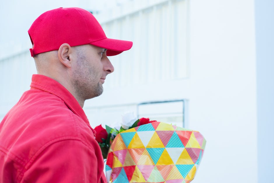 A delivery person wearing a red cap and red jacket is holding a vibrant floral gift wrapped in colorful geometric patterned paper, with a white ribbon accent. The flowers include red roses, white blos