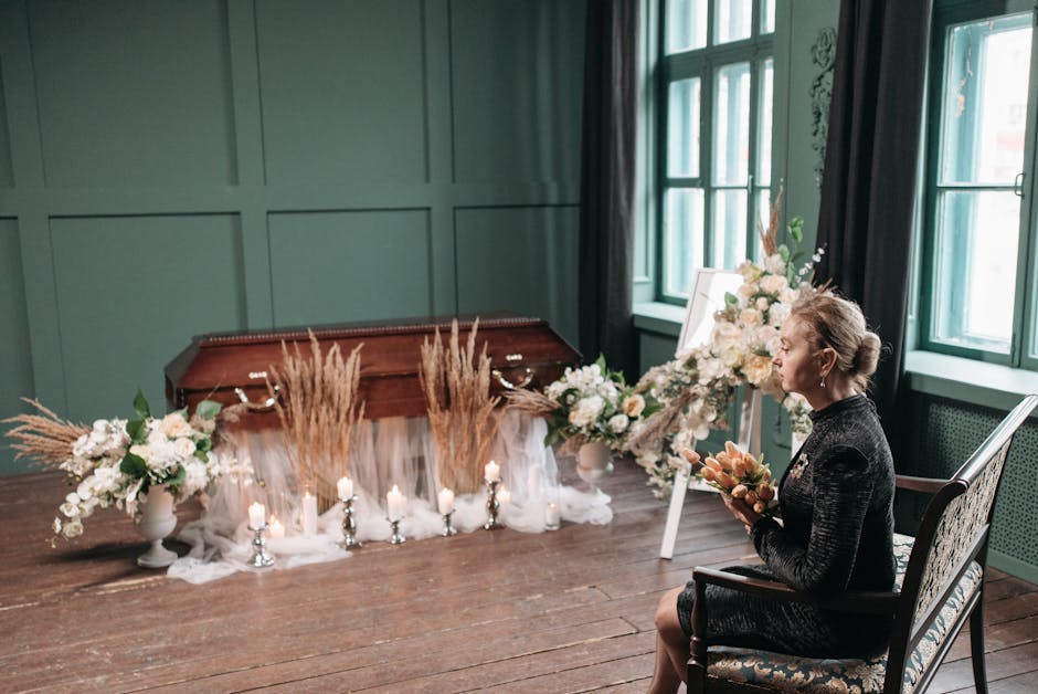 A woman with blonde hair styled in an updo, dressed in a black textured dress, sitting on an elegant wooden chair in a softly lit room with green paneled walls and large windows with dark curtains. Sh