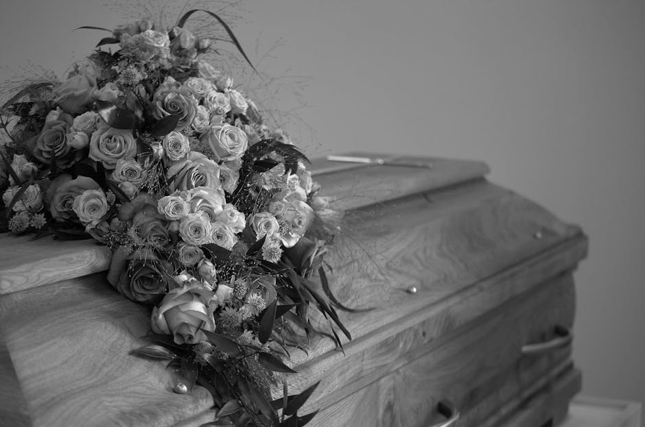A large, elegant floral arrangement of fresh roses and other mixed flowers in various sizes and shades, resting on top of a closed wooden casket. The bouquet features roses with tightly packed petals,