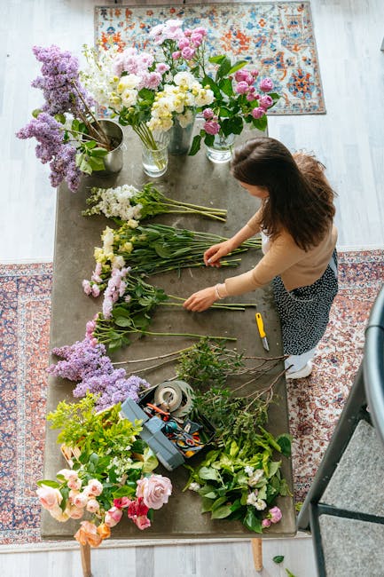 A woman with brown hair, dressed in a beige top and patterned skirt, arranging fresh flower stems on a rustic wooden table indoors. The table is covered with an array of colorful flowers, including pi