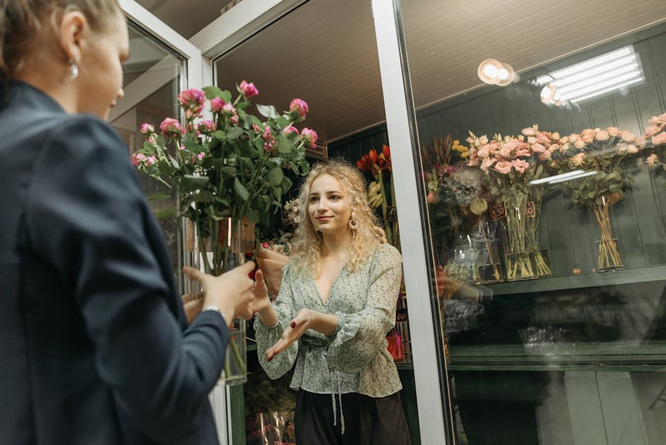 A young woman with curly blonde hair, wearing a light-colored, patterned blouse, is receiving a bouquet of fresh pink roses wrapped in brown paper from a staff member at flowerssouthgate.co.uk. The bo