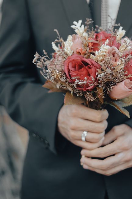 A close-up of a person in a dark suit holding a bouquet of fresh flowers, featuring large blush pink roses, white baby's breath, and dried foliage, wrapped in brown paper with a simple ribbon, set aga