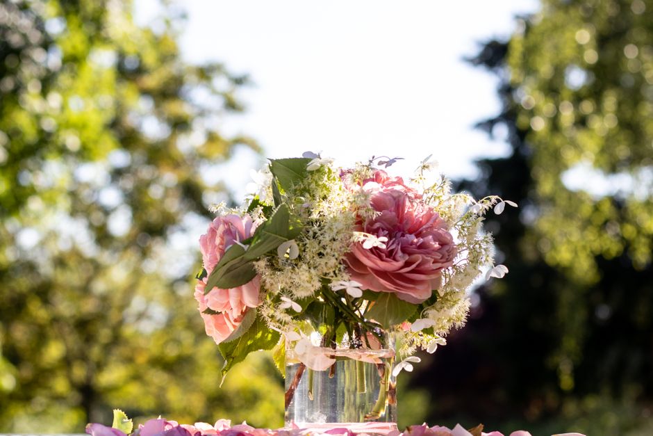 A floral arrangement featuring a bouquet of large, soft pink peonies with ruffled petals, accented by delicate white filler flowers and lush green leaves, all placed in a clear glass jar. The bouquet 