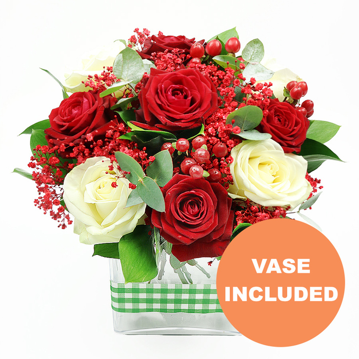 This detailed product image portrays a luxurious red and white rose arrangement crafted in Southgate, presented against a clean, bright white background that highlights every nuance of colour and texture. The central focus is a generous cluster of deep red roses, their petals plush and fully unfurled, paired with creamy white roses whose softly layered petals create a romantic, timeless feel. Scattered throughout the bouquet are fine sprays of tiny, vividly red filler flowers, reminiscent of gypsophila, that contribute a light, cloud-like texture and amplify the festive mood. Nestled between the blossoms, round, glossy red hypericum berries catch the eye, their polished surfaces reflecting the studio light and echoing the rich tones seen in seasonal décor along Southgate High Street during the winter months. Surrounding this interplay of blooms and berries, layers of fresh greenery offer a lush frame: dark, sturdy foliage provides structure and volume, while sprigs of eucalyptus, with their slightly bluish-green, oval leaves, lend a contemporary touch and suggest a subtle, refreshing fragrance. The stems are neatly grouped in a sleek, clear rectangular glass vase, perfectly proportioned to the arrangement and allowing a straightforward view of the cleanly cut stems inside. Around the vase, a green and white gingham ribbon is wrapped in a precise band, its country-style pattern bringing charm and warmth, as if the bouquet had been prepared for a cosy Southgate dining table or a welcoming hallway near Oakwood station. In the lower right corner of the image, a bold orange circular badge bearing the white uppercase text VASE INCLUDED confirms that this is a complete, ready-to-display gift-ideal for anniversaries, romantic surprises, festive entertaining, or simply brightening a North London living room with fresh flowers delivered from Flowers Southgate.